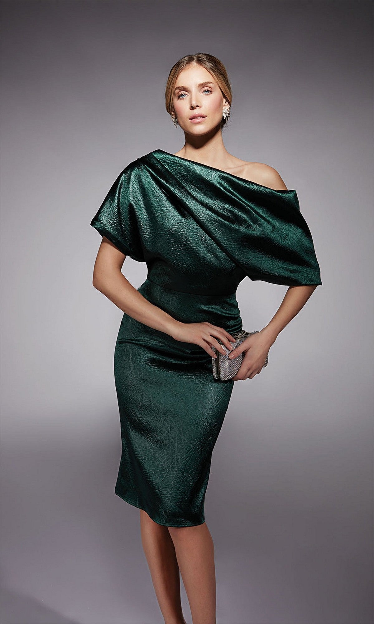A woman poses in the Alyce 70060 knee length semi-formal dress in metallic dark green textured charmeuse, holding a silver clutch against a plain gray background.